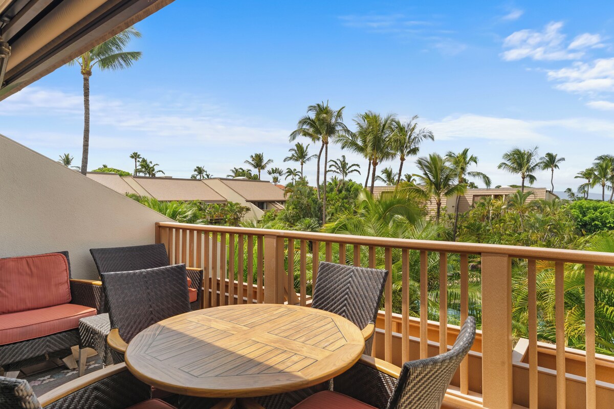 A balcony with outdoor furniture, including a round wooden table and wicker chairs, overlooking a tropical landscape with palm trees and a clear sky.