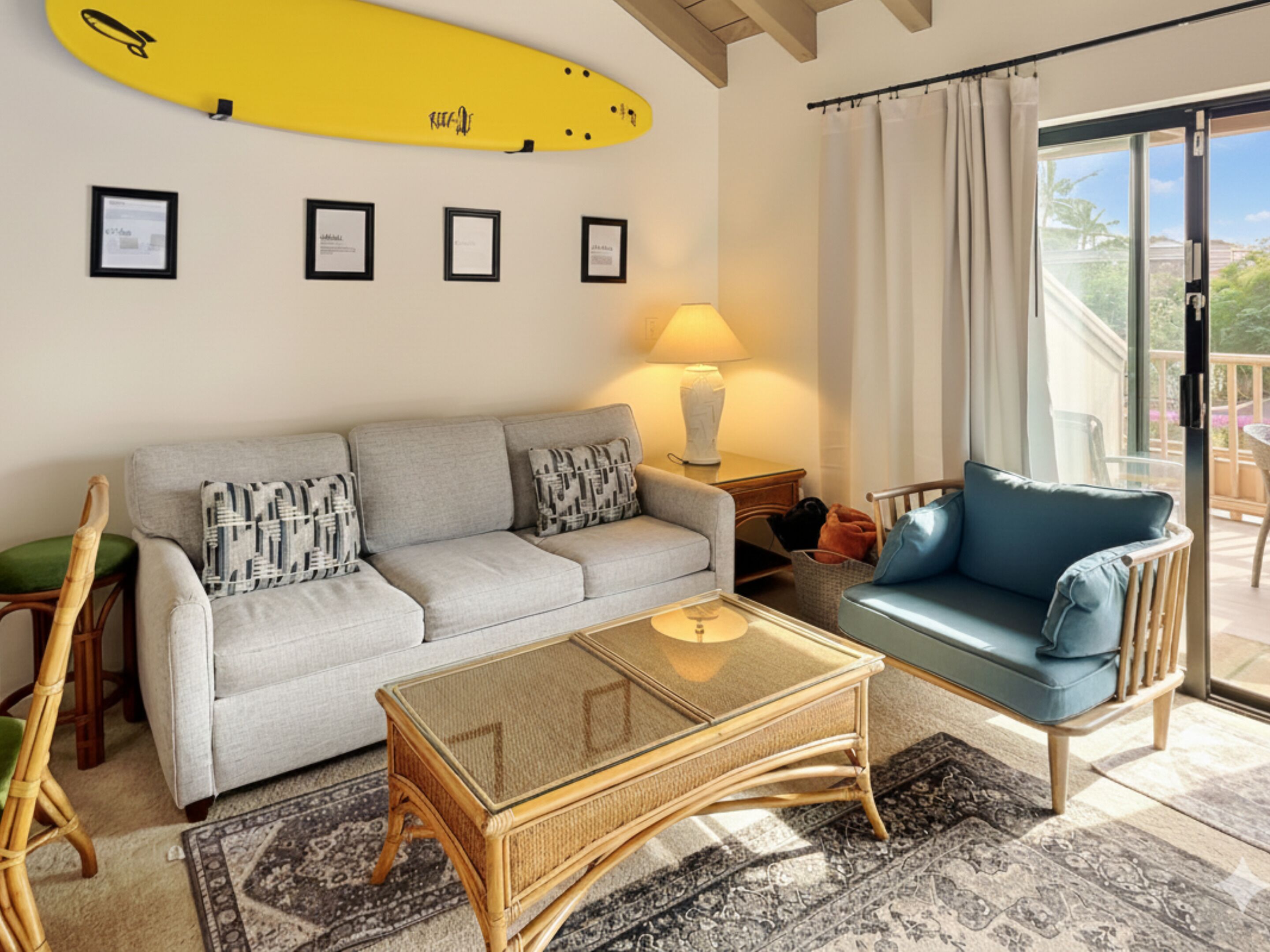 A living room with a sofa, a chair, a glass coffee table, a lamp, and a surfboard on the wall.