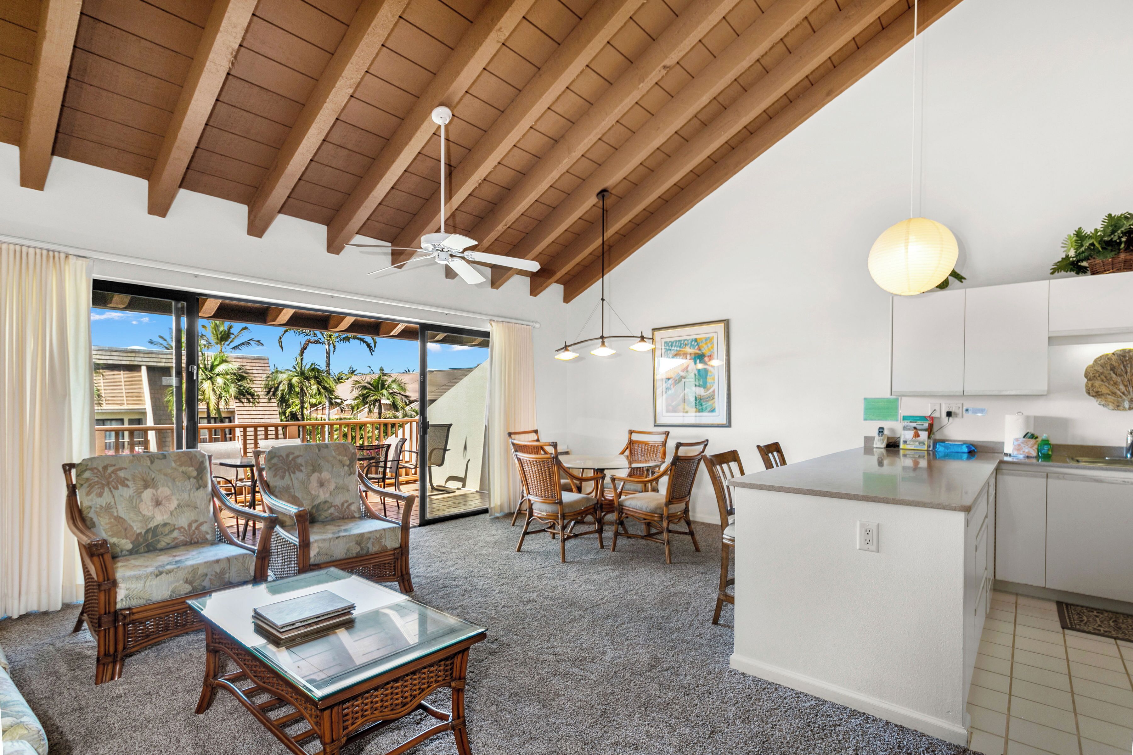 A spacious living area with a high ceiling, wooden beams, a glass coffee table, wicker furniture, a kitchen island, and a view of palm trees through large sliding doors.
