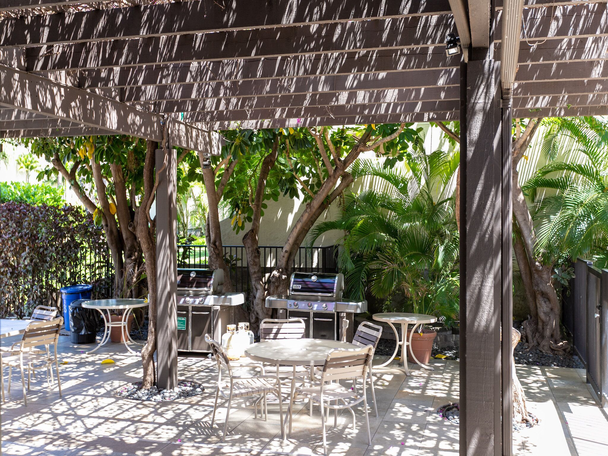 An outdoor patio area with tables and chairs, a barbecue grill, and a pergola providing shade.