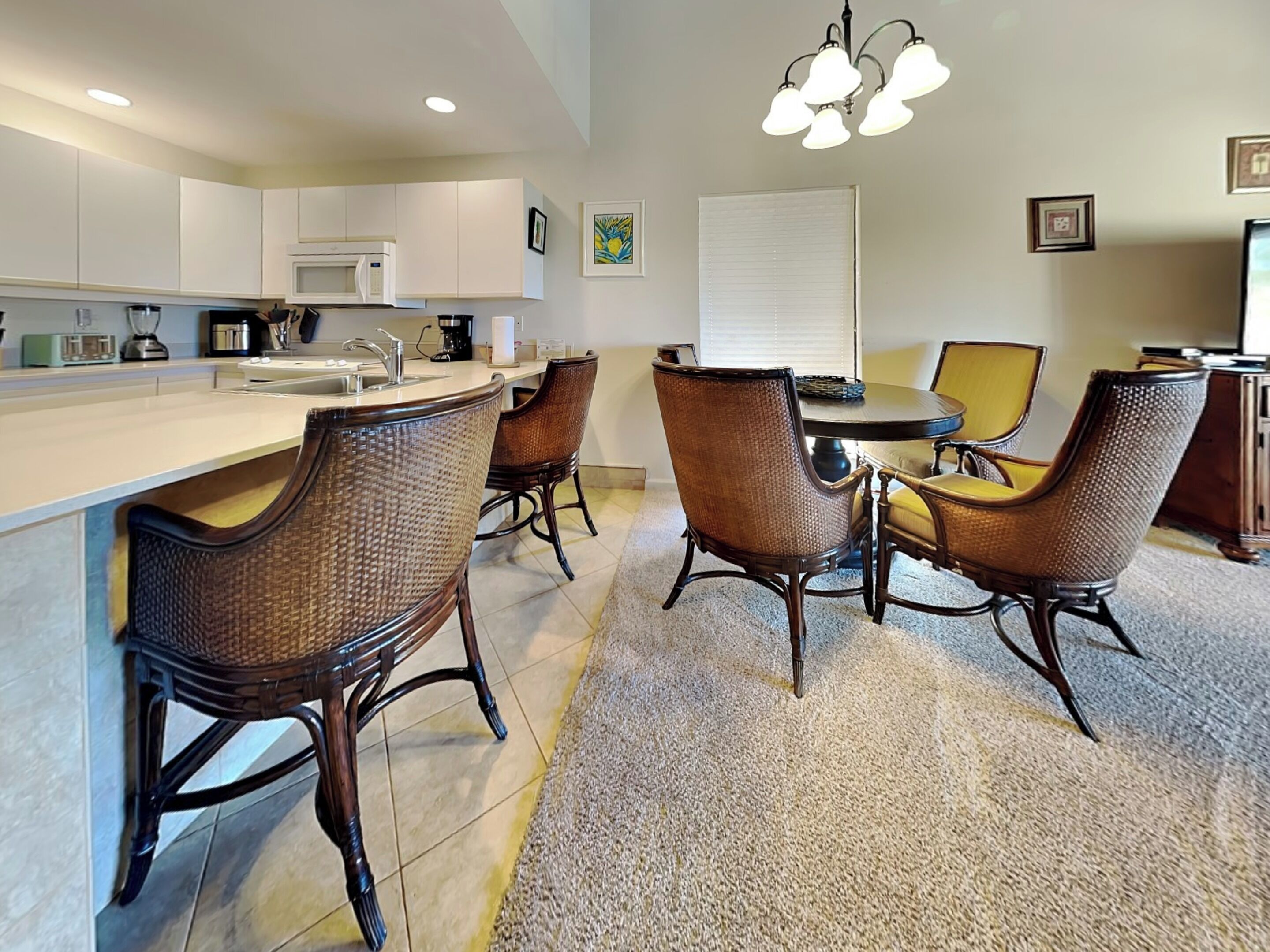 A modern kitchen with white cabinets, a microwave, and a sink. There are wicker chairs around a dining table with a yellow cushion. A chandelier hangs above the table.