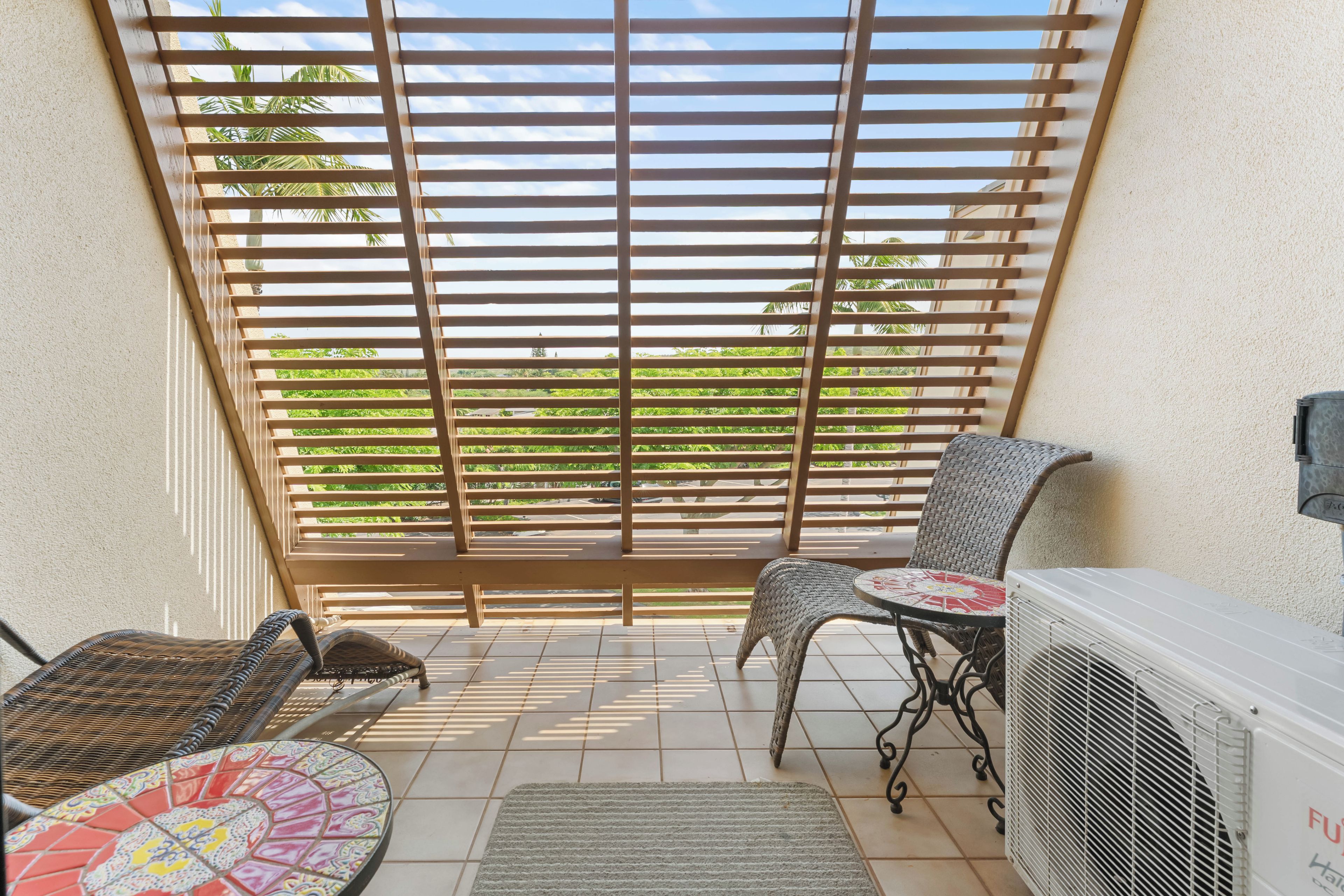 A sunlit balcony with a view of greenery, a wicker chair, a small table with a patterned tablecloth, and a white air conditioning unit.