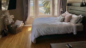 Un dormitorio con cama, televisor montado en la pared, ventana con vista a la playa y mesita de noche con control remoto.