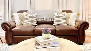 A brown leather sofa with decorative pillows and a small table with books and a vase of flowers. - Cozy Getaway | Splendid Historic Thibodaux Gem (Thibodaux)