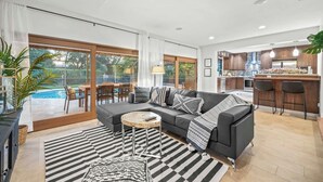 A modern living room with a grey sofa, a round coffee table, and a striped rug. There is a large window with a view of a pool and greenery. The kitchen has wooden cabinets and a stone backsplash. - Family Beach Villa w/ Home Theater & Heated Pool! (Hollywood)