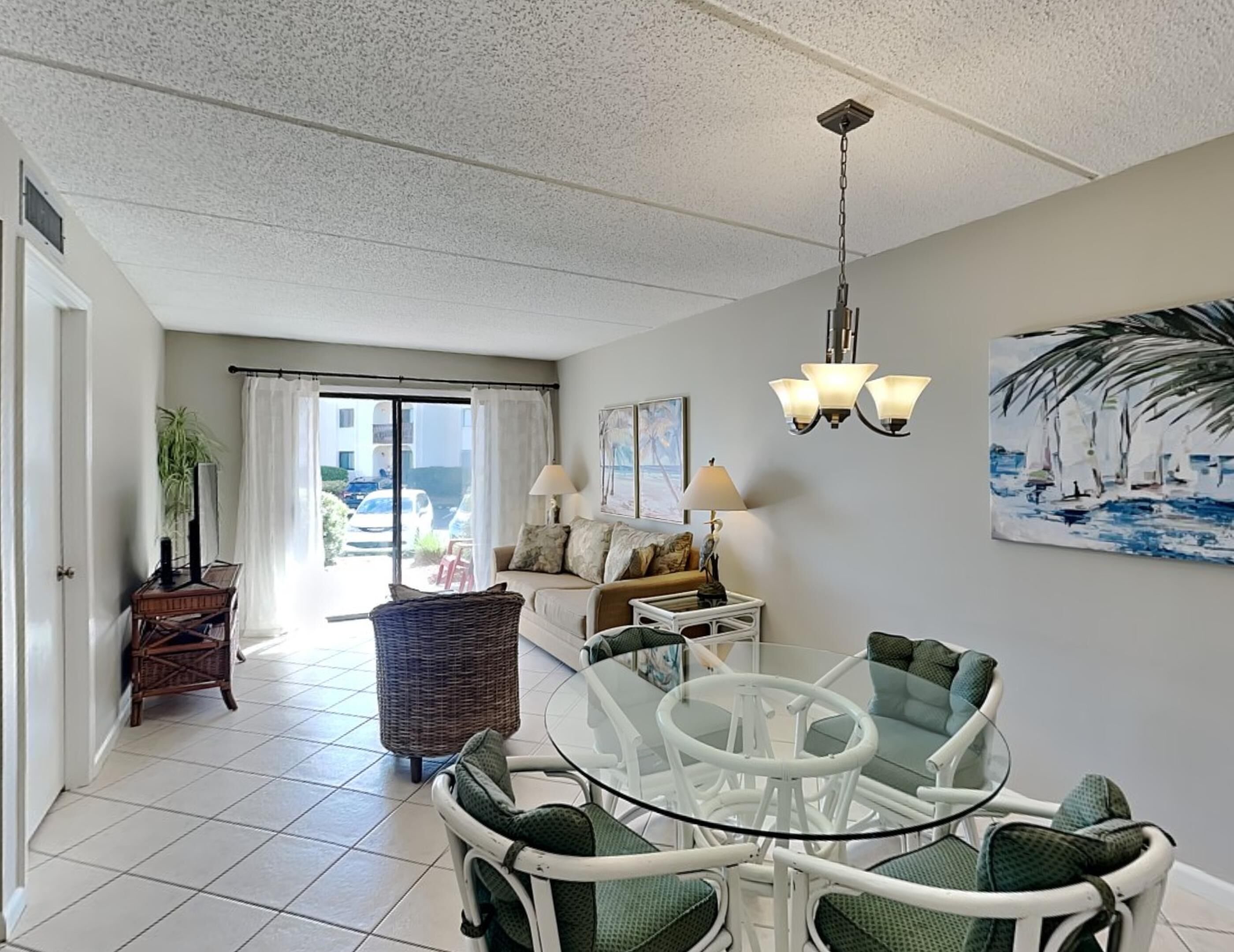 A spacious living area with a glass dining table surrounded by chairs, a sofa, and a wicker chair. A painting of a beach scene adorns the wall, and a ceiling light fixture is visible.