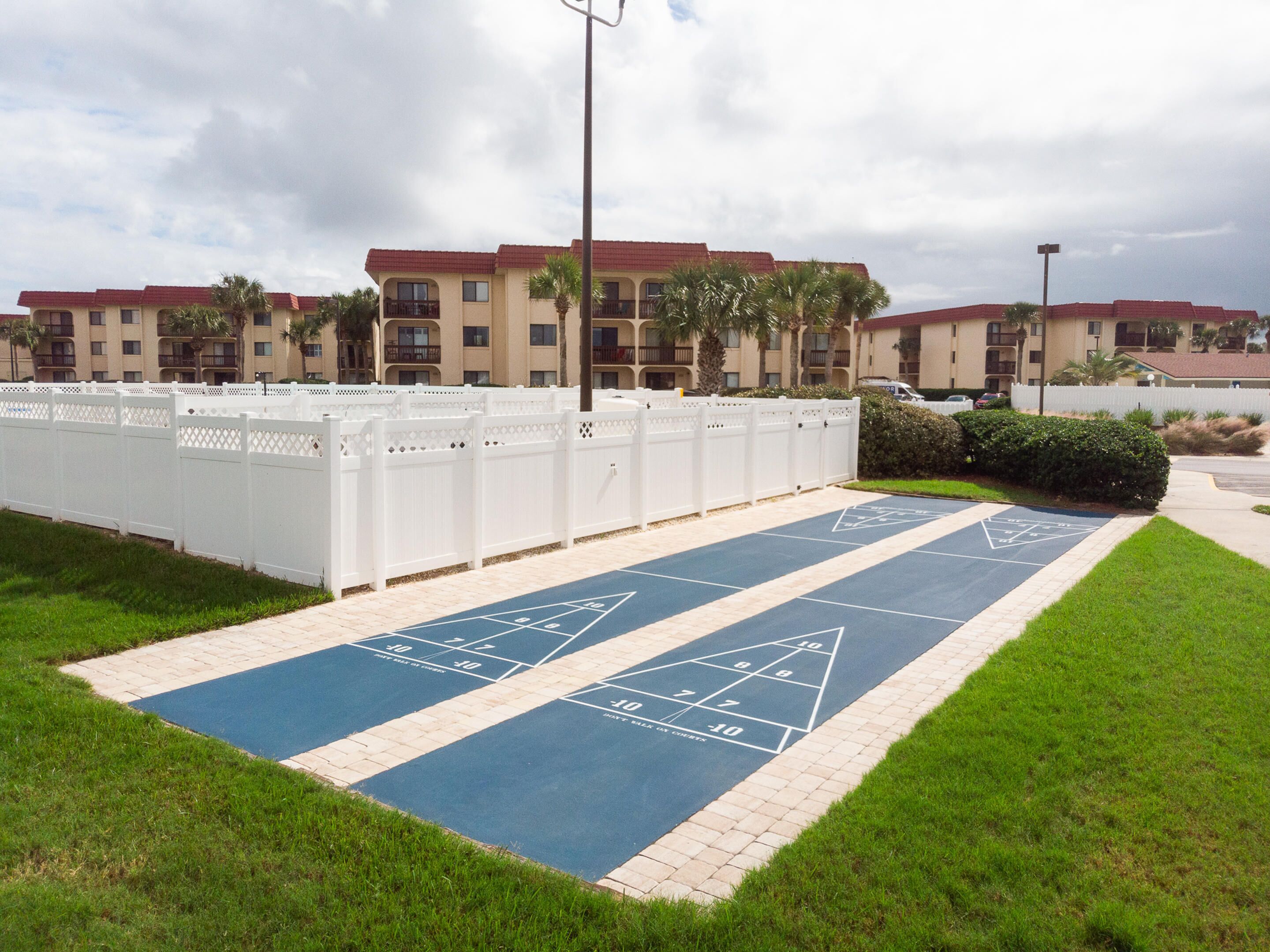 A shuffleboard court with a white fence and a building in the background.