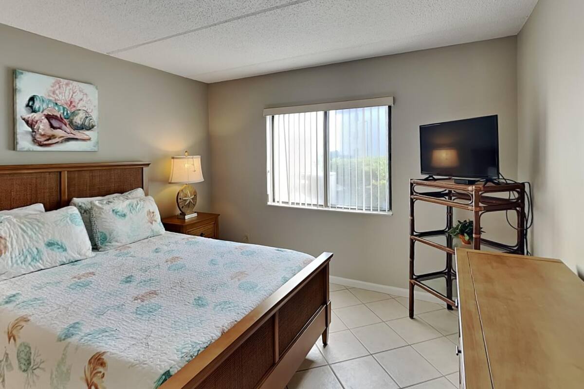 Un dormitorio con cama, televisor sobre un mueble, ventana con persianas y un cuadro en la pared.