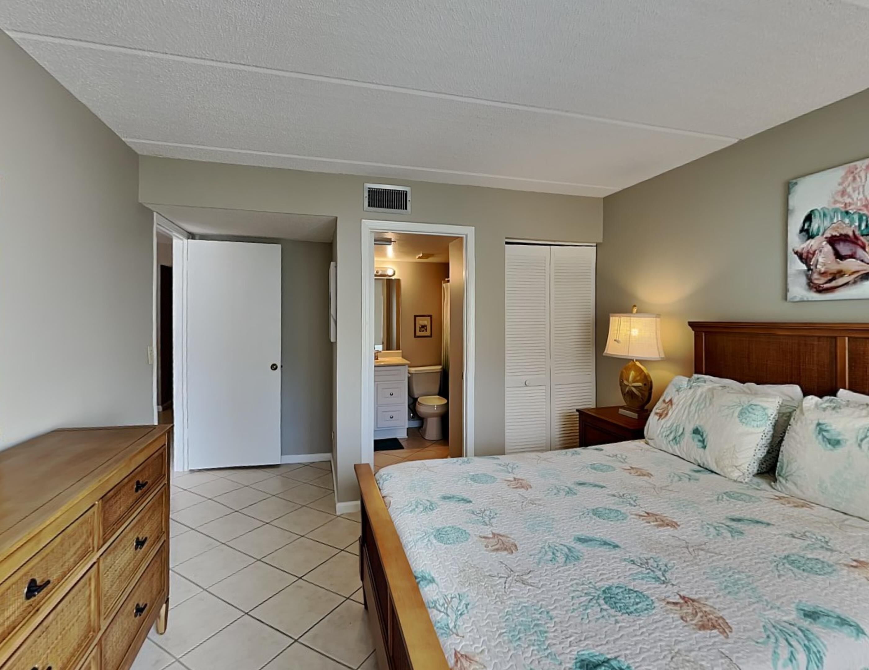 Une chambre à coucher avec un lit, une commode en bois, une table de chevet avec une lampe, un placard et une salle de bain visible à travers une porte ouverte.