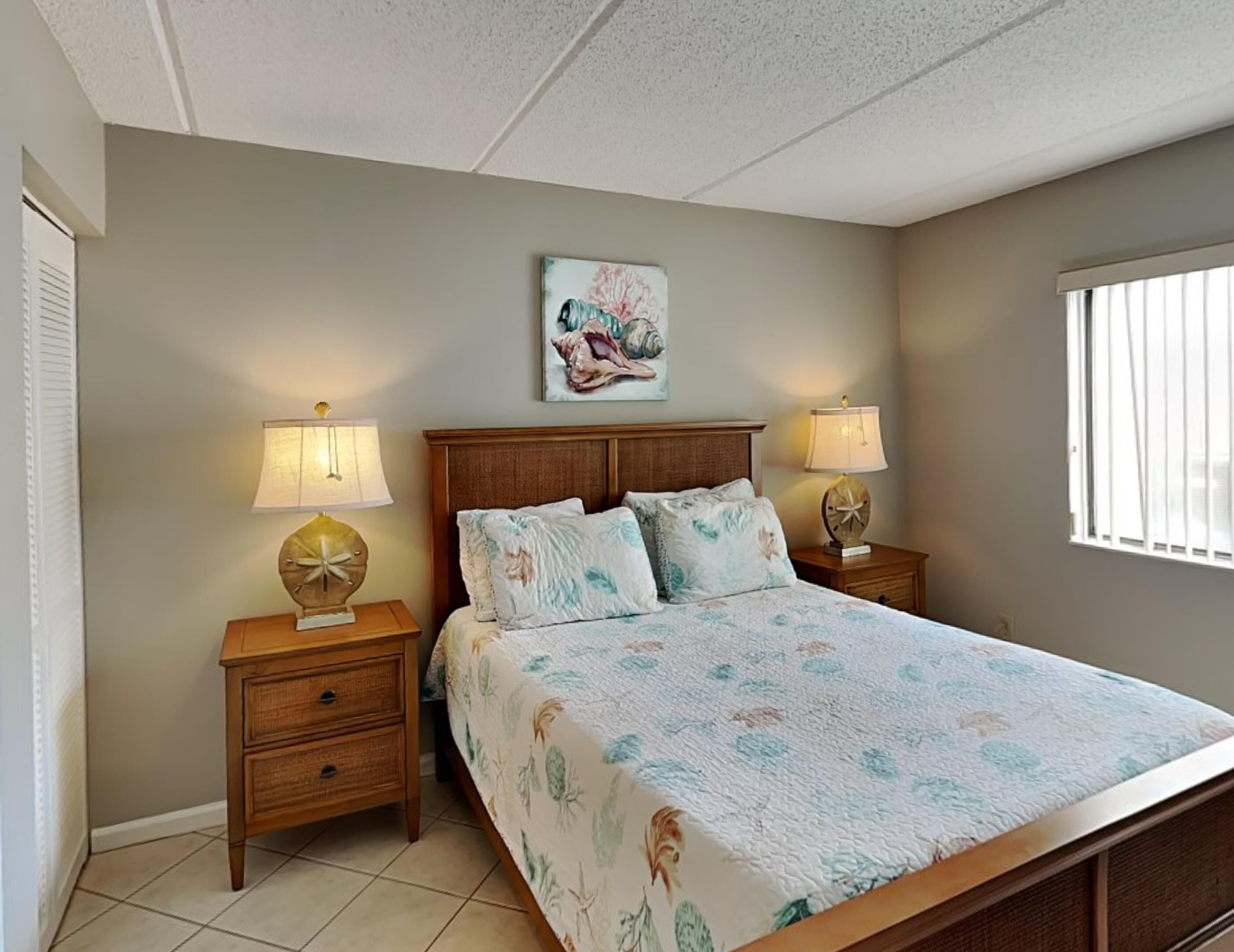A bedroom with a bed, two bedside tables with lamps, a wall-mounted painting, and a window.