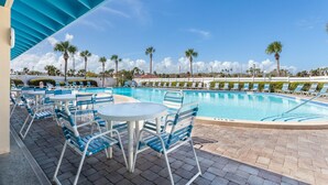 A pool area with lounge chairs and tables, surrounded by palm trees and a clear sky.