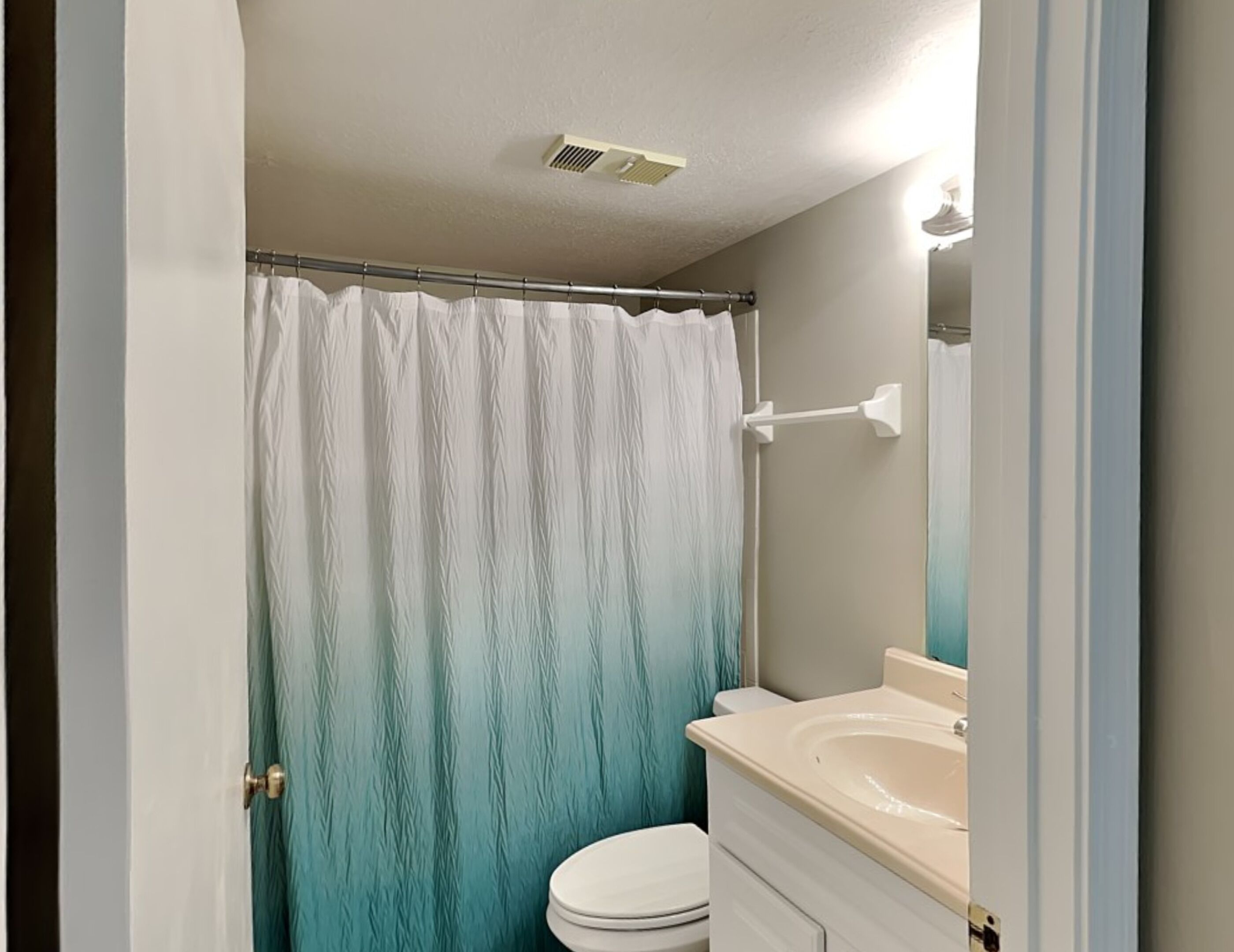 Un baño con un inodoro blanco, un lavamanos color crema y una cortina de ducha color verde azulado.