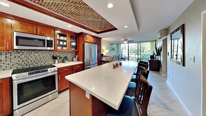 A modern kitchen with wooden cabinets, stainless steel appliances, a large island, and a dining area with chairs. - Pacific Shores by Maui Rental Group (Kihei)