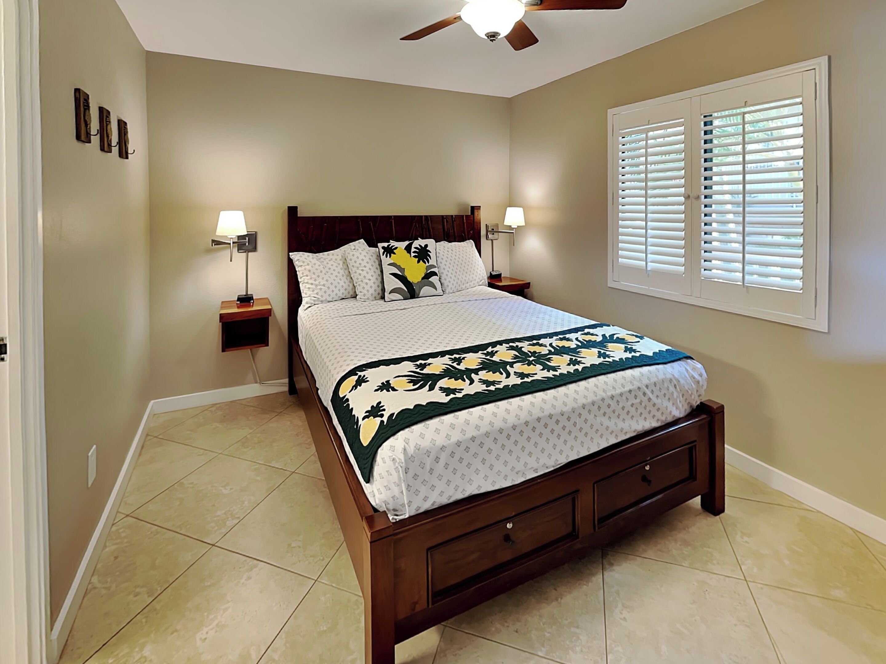 A bedroom with a bed, two bedside tables, a ceiling fan, a window with blinds, and a wall-mounted light fixture.