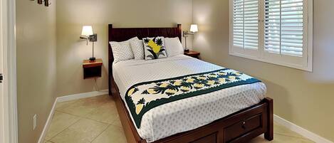 A bedroom with a bed, two bedside tables, a ceiling fan, a window with blinds, and a wall-mounted light fixture.