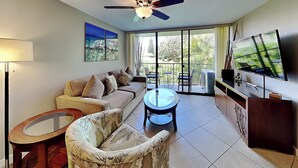 A living room with a sofa, a glass coffee table, a patterned armchair, a ceiling fan, a TV, and a balcony with outdoor furniture. - Pacific Shores by Maui Rental Group (Kihei)