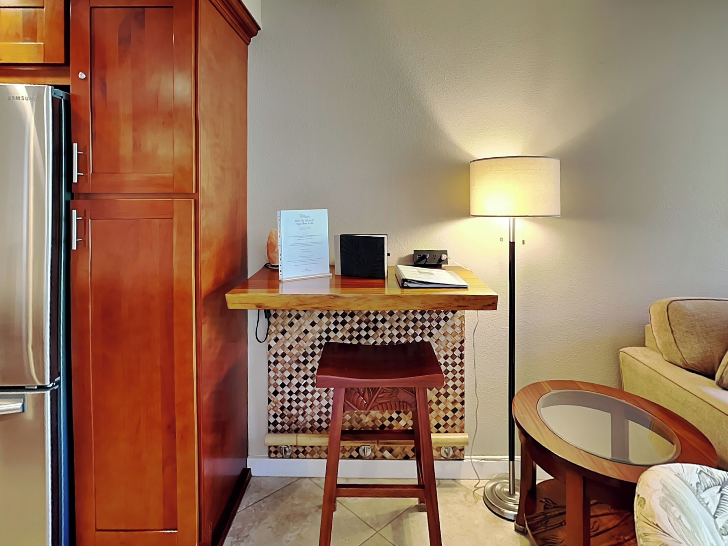 A compact kitchen with a refrigerator, a wooden countertop, a small stool, a table with a lamp, and a patterned backsplash.