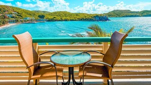 A balcony with wicker chairs and a glass table overlooking a turquoise sea, with hills and buildings in the background. - Stunning St. Thomas Condo for Your Tropical Escape (St. Thomas)