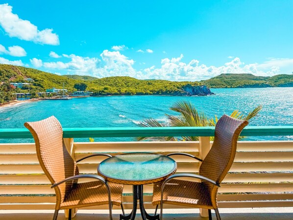 A balcony with wicker chairs and a small table overlooking a turquoise sea and green hills.