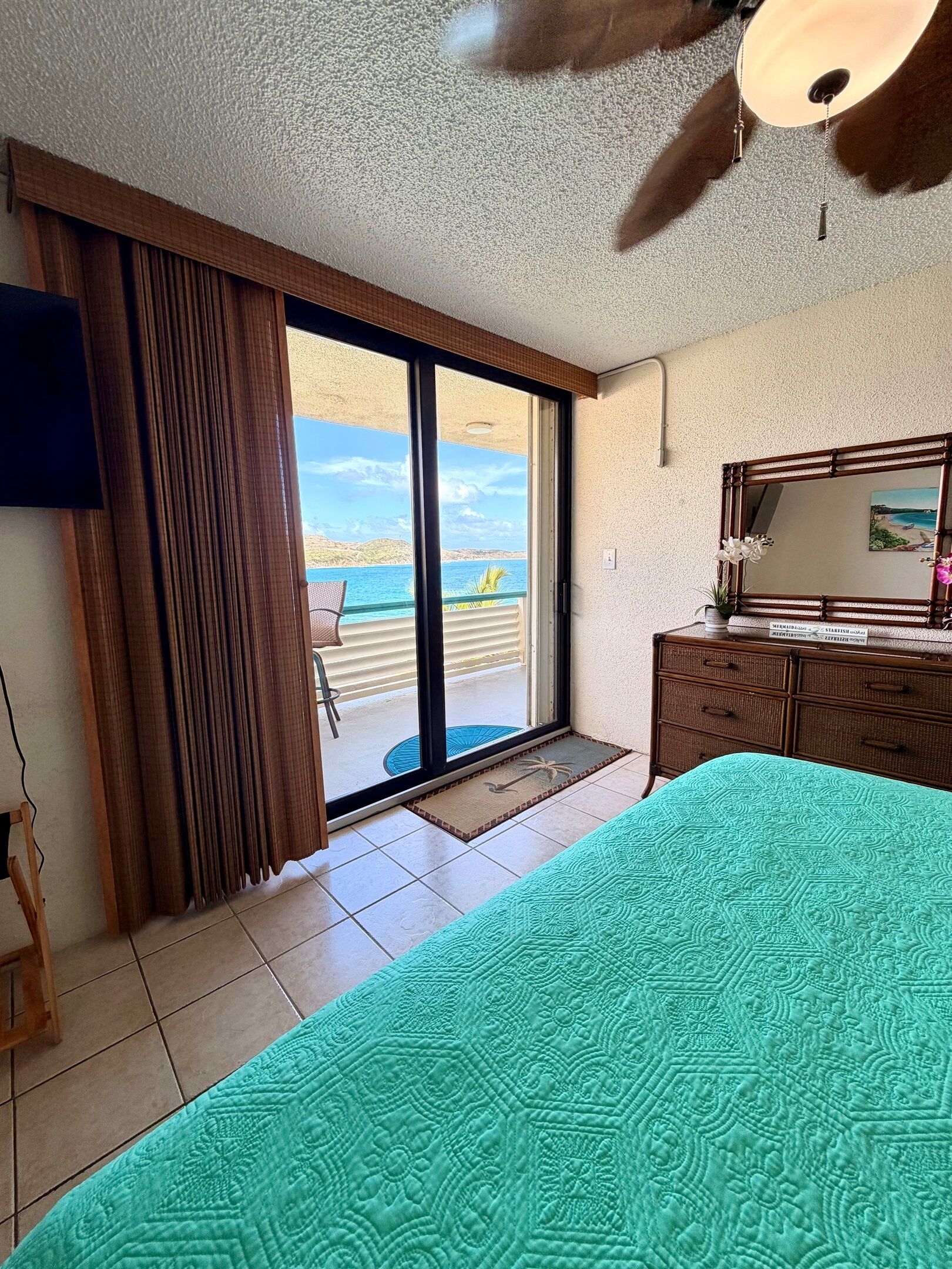 A hotel room with a bed, a ceiling fan, a large window with curtains, a view of the sea, and a wooden dresser.