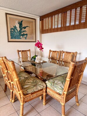 A dining area with a glass-top table surrounded by intricately carved bamboo chairs, a framed artwork on the wall, and a bamboo screen.