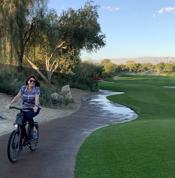 Stroll through a lush landscape along a winding pathway surrounded by trees and shrubs, with a golf course visible in the distance.