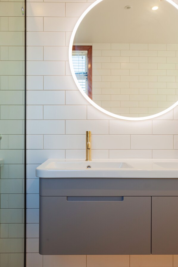 A bathroom with a white tiled wall, a round mirror with a light, and a white sink with a gold faucet. - Wigram Hotel (Christchurch)