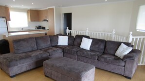 A modern living room with a large sectional sofa, a matching ottoman, and a kitchen area with wooden cabinets and a window.