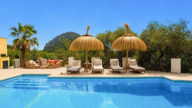 A swimming pool with lounge chairs and thatched umbrellas, surrounded by greenery and a mountain in the background.
