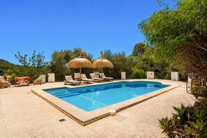 Une piscine rectangulaire, bordée d’une terrasse carrelée, entourée de chaises longues et de parasols, avec une végétation luxuriante en arrière-plan.