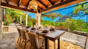 An outdoor dining area with a stone floor, wooden table, wicker chairs, and a view of greenery and mountains.