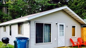 A single-story house with a gabled roof, a front porch with a red chair, and a barbecue grill. - Cabin 2 | Headwater (Nevis Township)