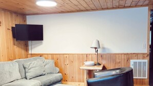 A cozy living room with a grey sectional sofa, two black leather ottomans, a small white table with a lamp, and a mounted TV.