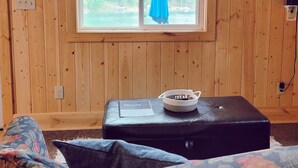 A room with wooden walls, a window with a view of trees, a television mounted on the wall, and a black ottoman with a welcome card. - Cabin 4 | Moonbeam (Nevis Township)