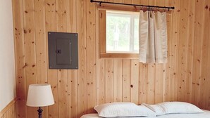 A wooden-paneled room with a bed, a nightstand, a lamp, a window with curtains, and a wall-mounted electrical box. - Cabin 4 | Moonbeam (Nevis Township)