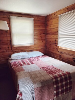 A wooden-paneled room with a bed, two windows with blinds, and a wall-mounted lamp. - Cabin 6 | Voyageur (Nevis Township)