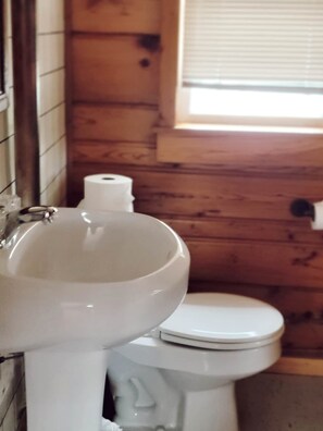 A bathroom with a white sink and toilet, wooden walls, and a window. - Cabin 6 | Voyageur (Nevis Township)