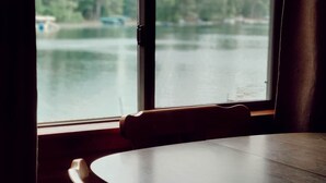 A room with a round table and a single wooden chair, a window with a view of a lake and trees.