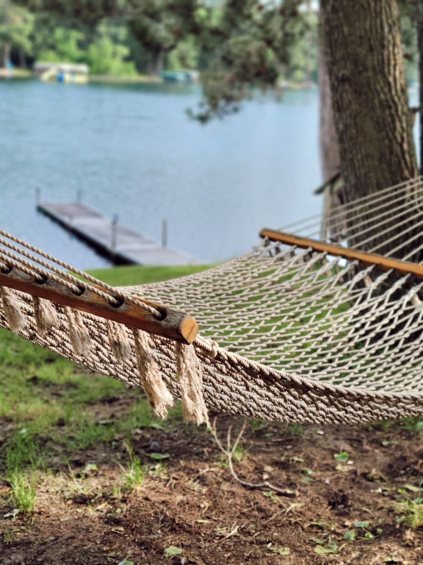 A hammock tied between trees by a lake with a dock in the background.