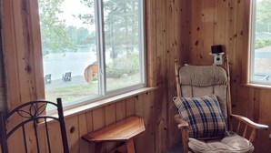 A wooden cabin with a large window overlooking a lake, a wooden bench, and a chair with a plaid cushion. - Cabin 8 | Northwoods (Nevis Township)