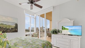 A bedroom with a bed, a TV, a ceiling fan, and a balcony with a view of the city.