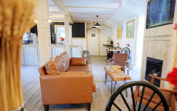Un salon chaleureux avec un canapé, une table basse en bois et une cheminée. Au fond, on aperçoit un coin cuisine avec une table à manger et des chaises.