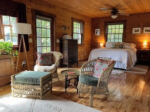 Ein gemütliches Blockhaus-Interieur mit Holzboden, einem Bett mit weißer Bettwäsche, Korbsesseln mit gemusterten Kissen, einem kleinen runden Tisch und einem Deckenventilator.
