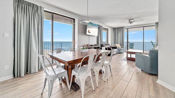 Uma sala de estar moderna com uma mesa de jantar de madeira, cadeiras brancas, um sofá cinza e uma TV fixada na parede.