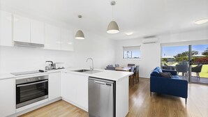 A modern kitchen with white cabinets, a dishwasher, and a coffee maker. There is a dining area with a blue sofa and a view of the outdoors. - Top Town Terrace - short walk to town center (Woolgoolga)