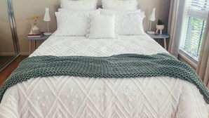 A neatly made bed with white bedding and a green knitted throw, a bedside table with a lamp, and a small plant.