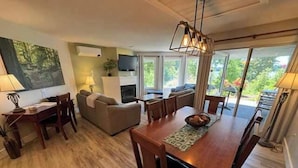 Uma sala de estar moderna com área de jantar, lareira e uma grande janela com vista para o verde.