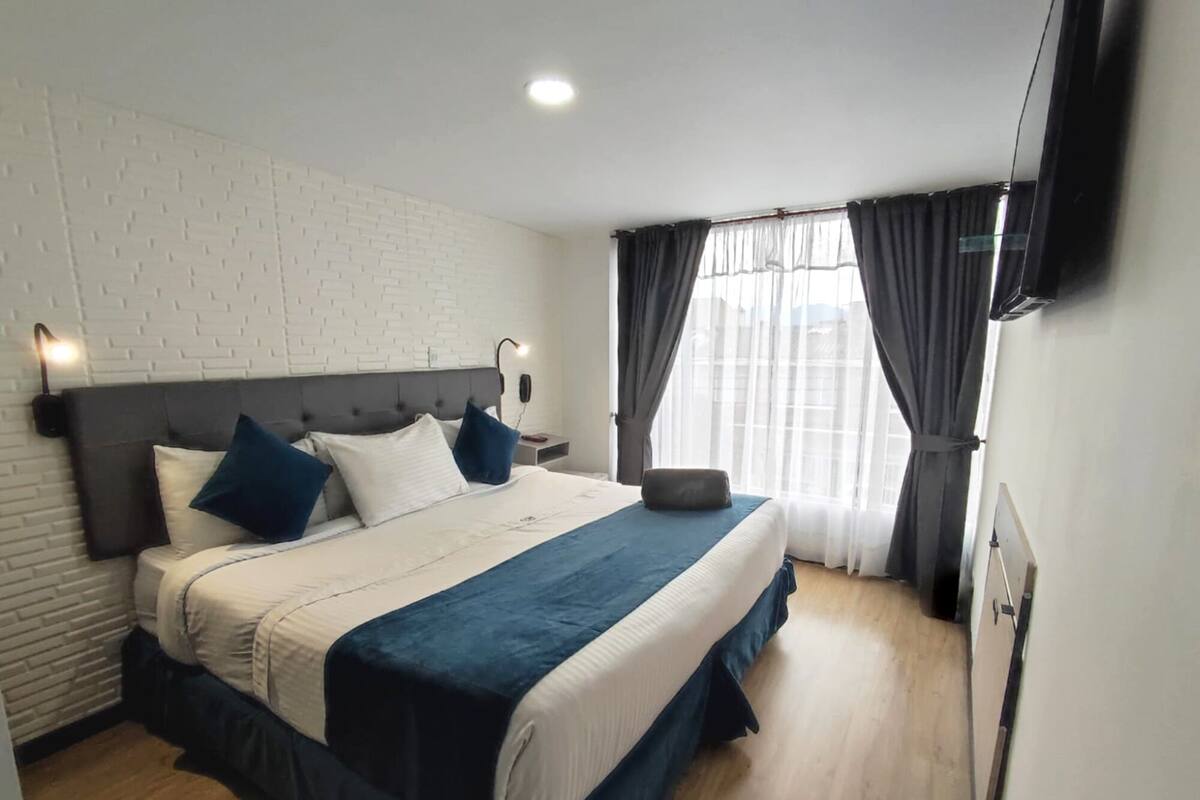 Une chambre d’hôtel avec un lit, une table de chevet, une lampe, une télévision et une fenêtre avec des rideaux.