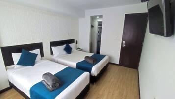 Quarto de hotel com duas camas, cada uma com colcha azul e branca, piso de madeira, parede de tijolos brancos e uma porta com uma televisão fixada.