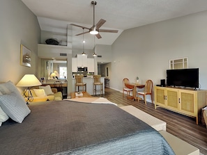 A bedroom with a large bed, a television, a dining area, and a kitchen in the background.